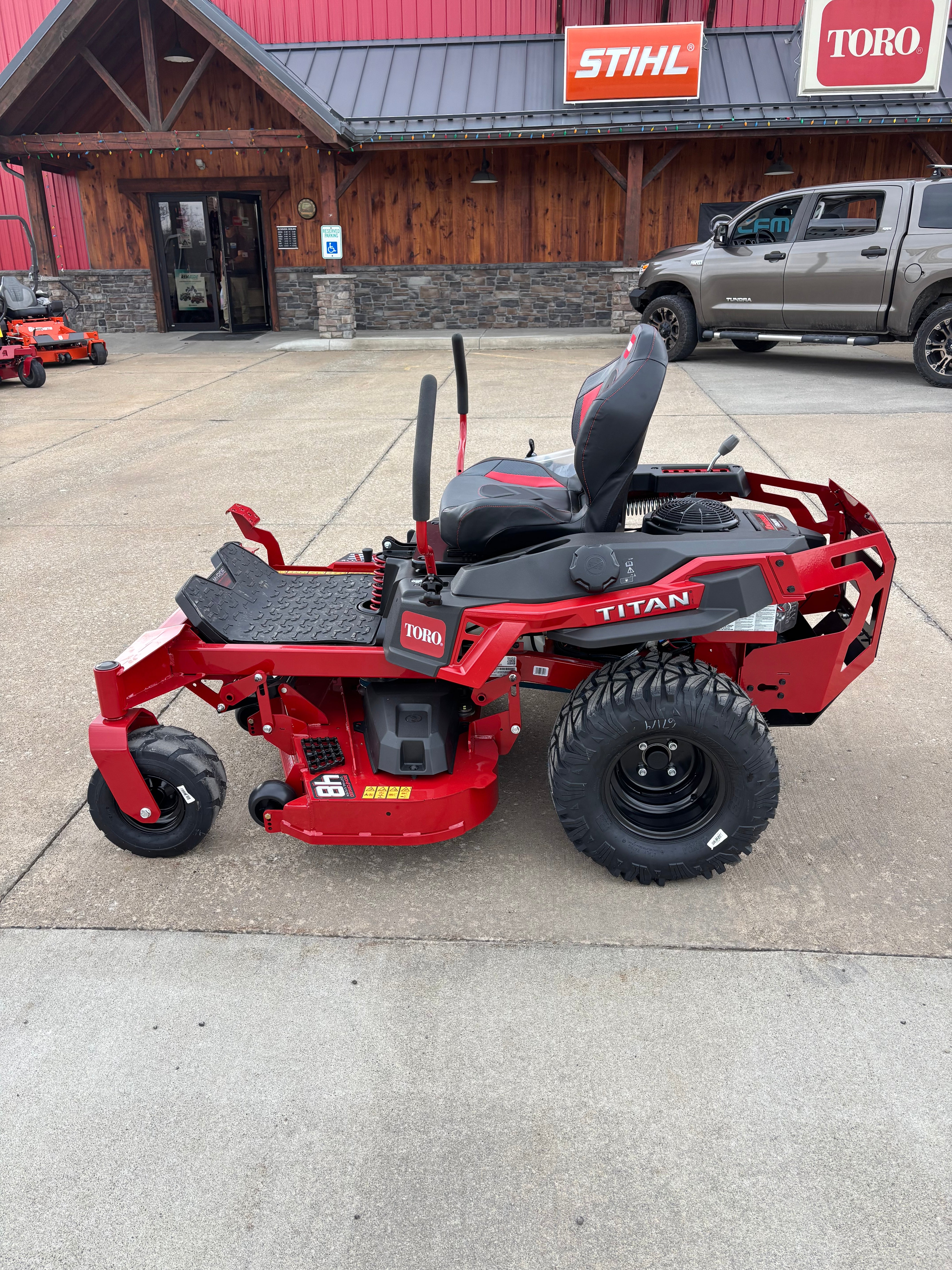 Red riding lawn mower on a concrete surface with a building and vehicles in the background.